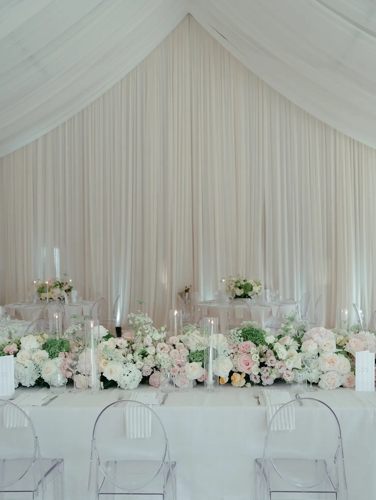 Elegant wedding reception table with white linens, blush floral arrangements, clear chairs, and sheer white drapery backdrop.