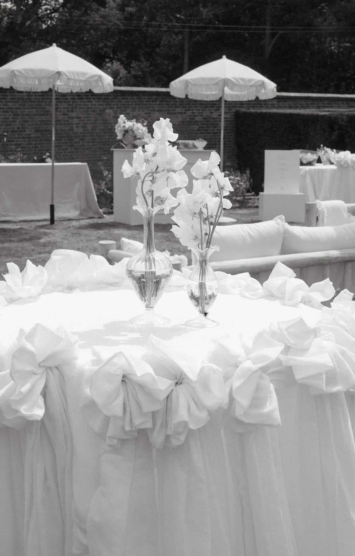 Outdoor wedding table with white draped cloth and two glass vases holding white flowers.