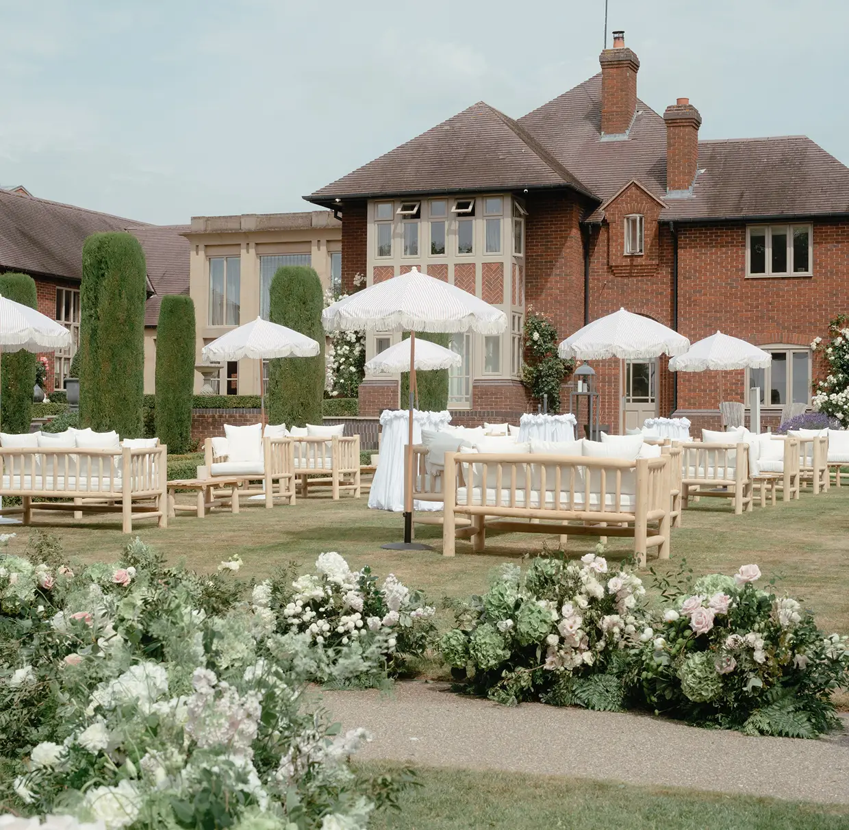 Outdoor wedding seating area with white umbrellas, cushioned wooden benches, and floral arrangements.