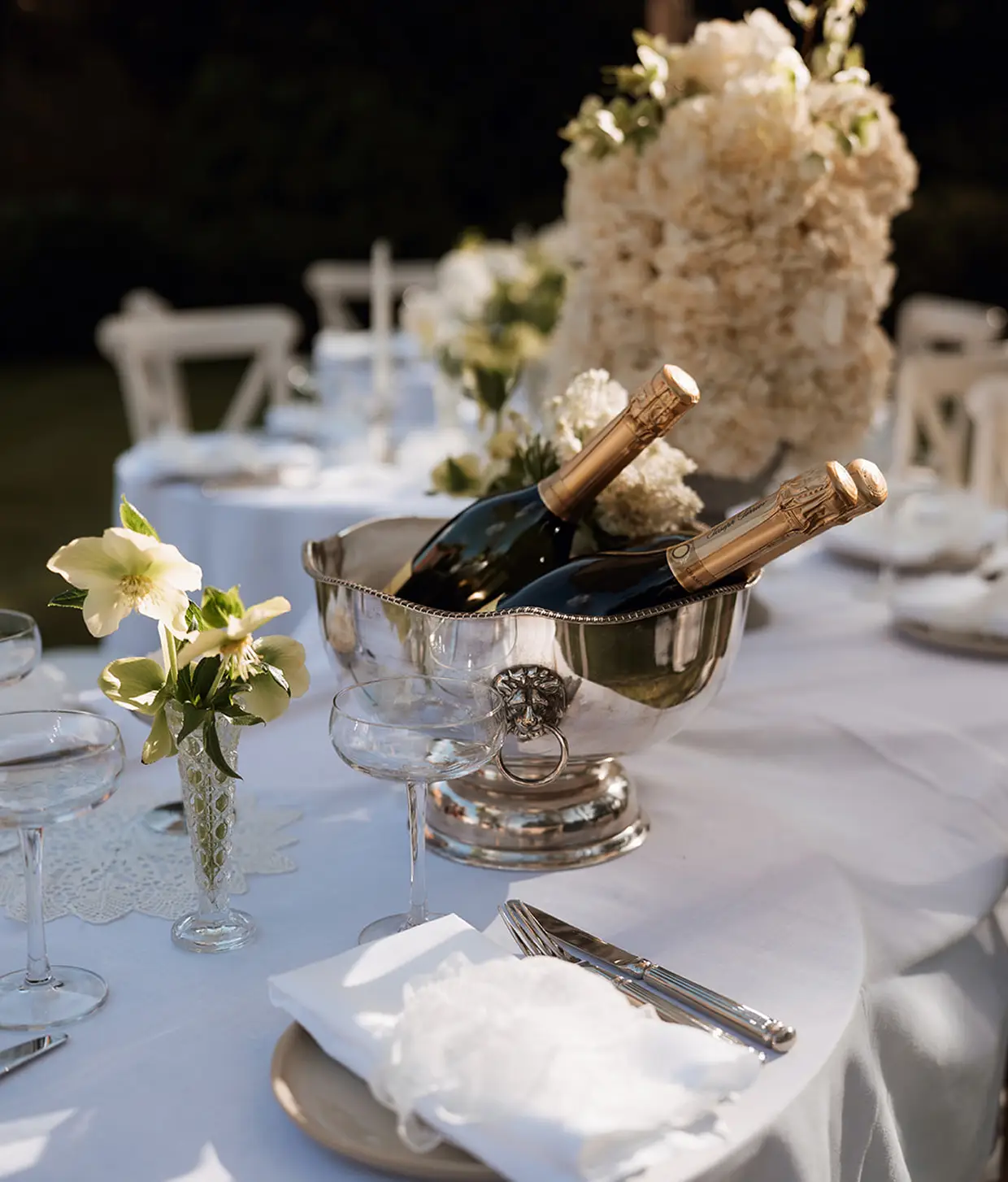 Two bottles of champagne in a silver ice bucket on an elegantly set white table with flowers and glassware.