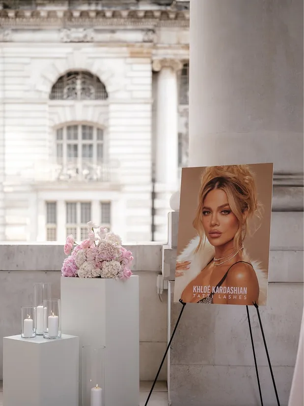 Portrait poster of Khloe Kardashian promoting Tati Lashes on an easel beside floral arrangements and candles outdoors.