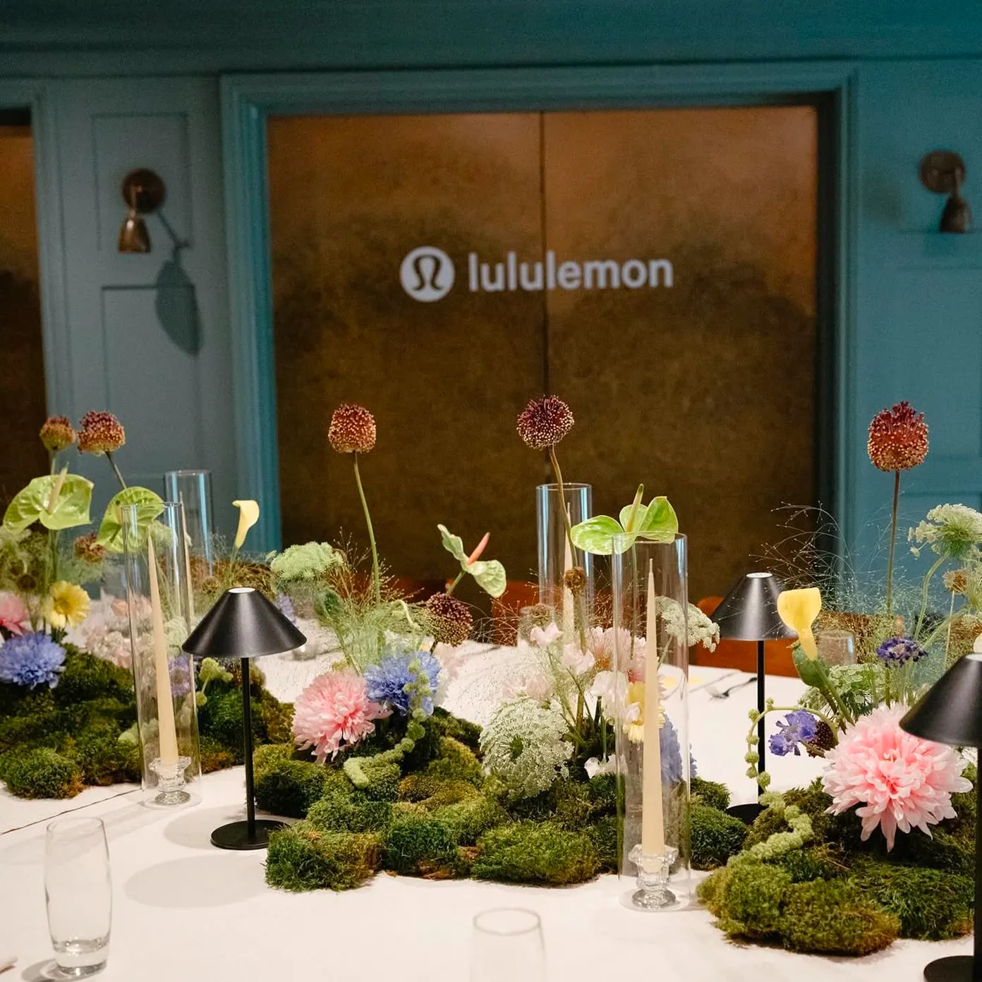 Table centerpiece with moss, colorful flowers, candles in glass holders, and black lamps under Lululemon sign.