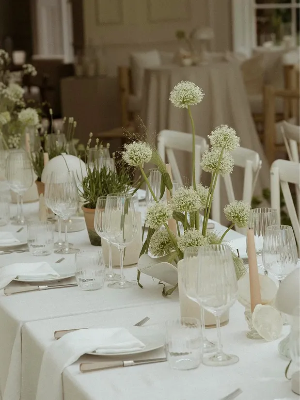 Elegant table setting with white tablecloth, cluster of white flowers in a vase, glassware, and pale candles.