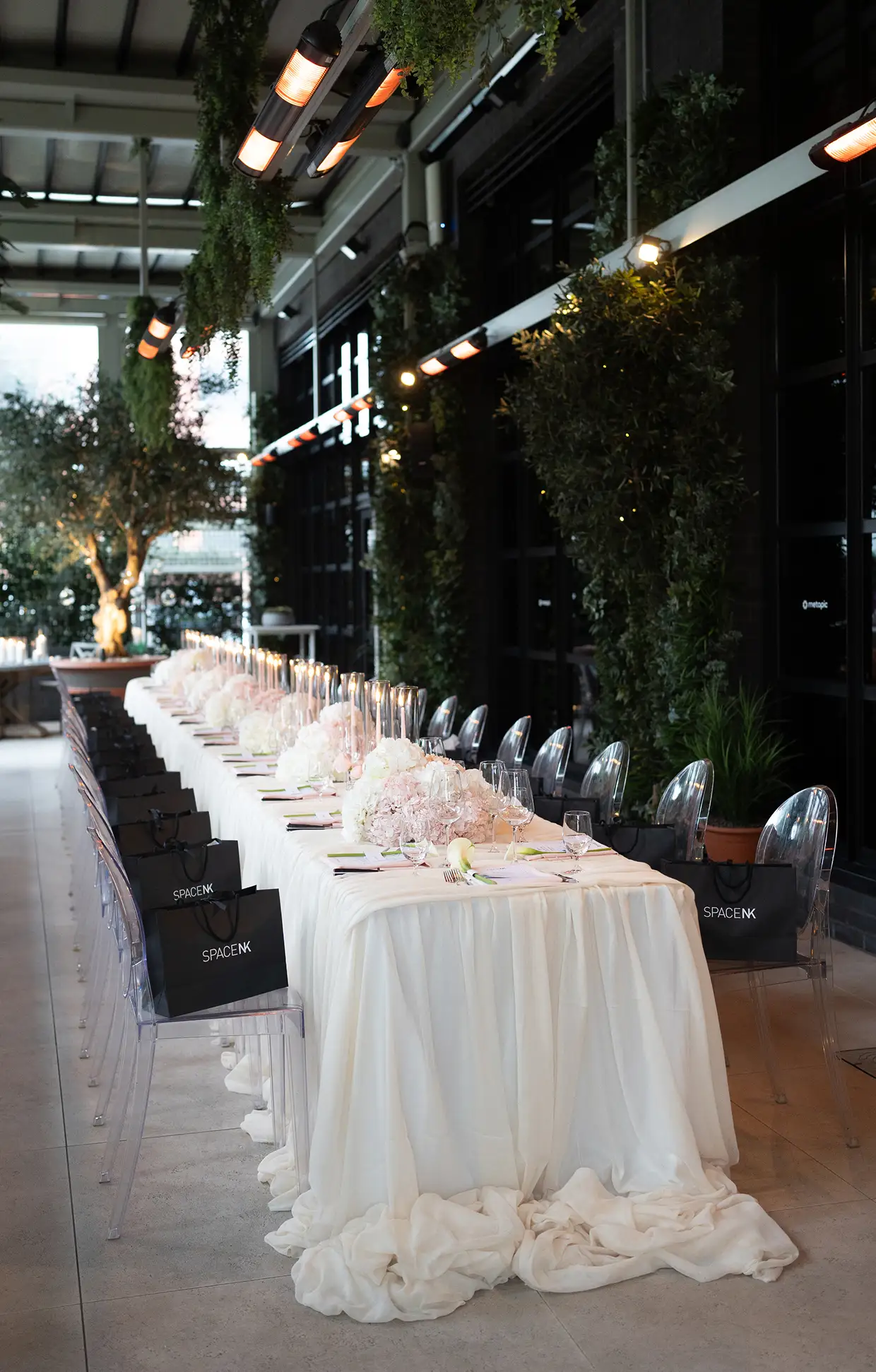 Long dining table with white tablecloth, floral centerpieces, candles, and transparent chairs with black SpaceNK bags.