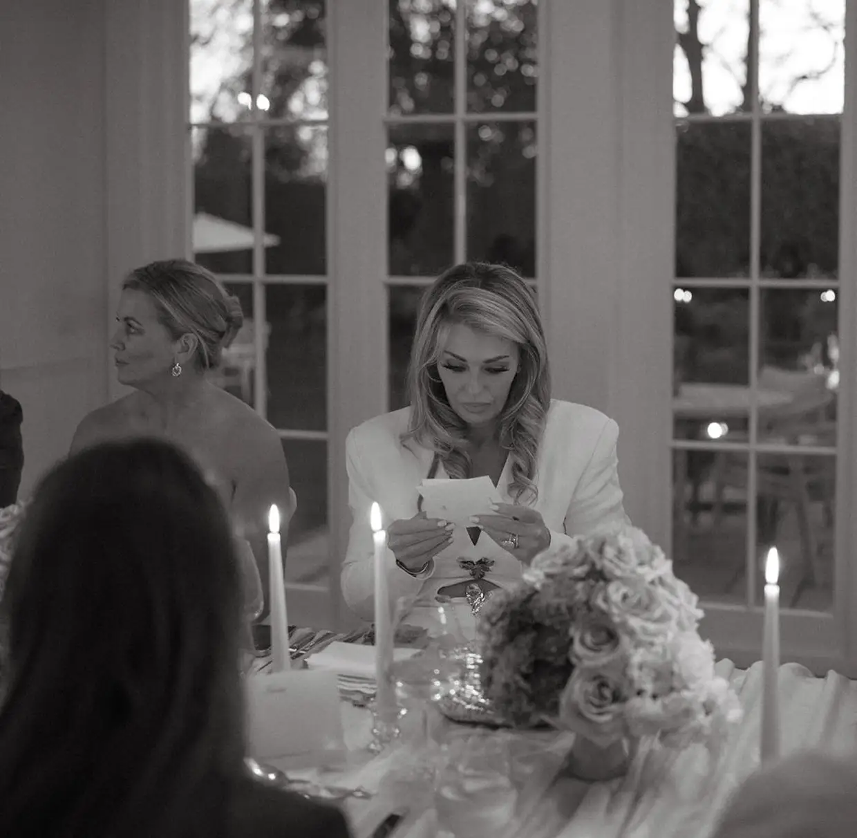 Woman in white jacket reading a note at a candlelit table with floral centerpiece.