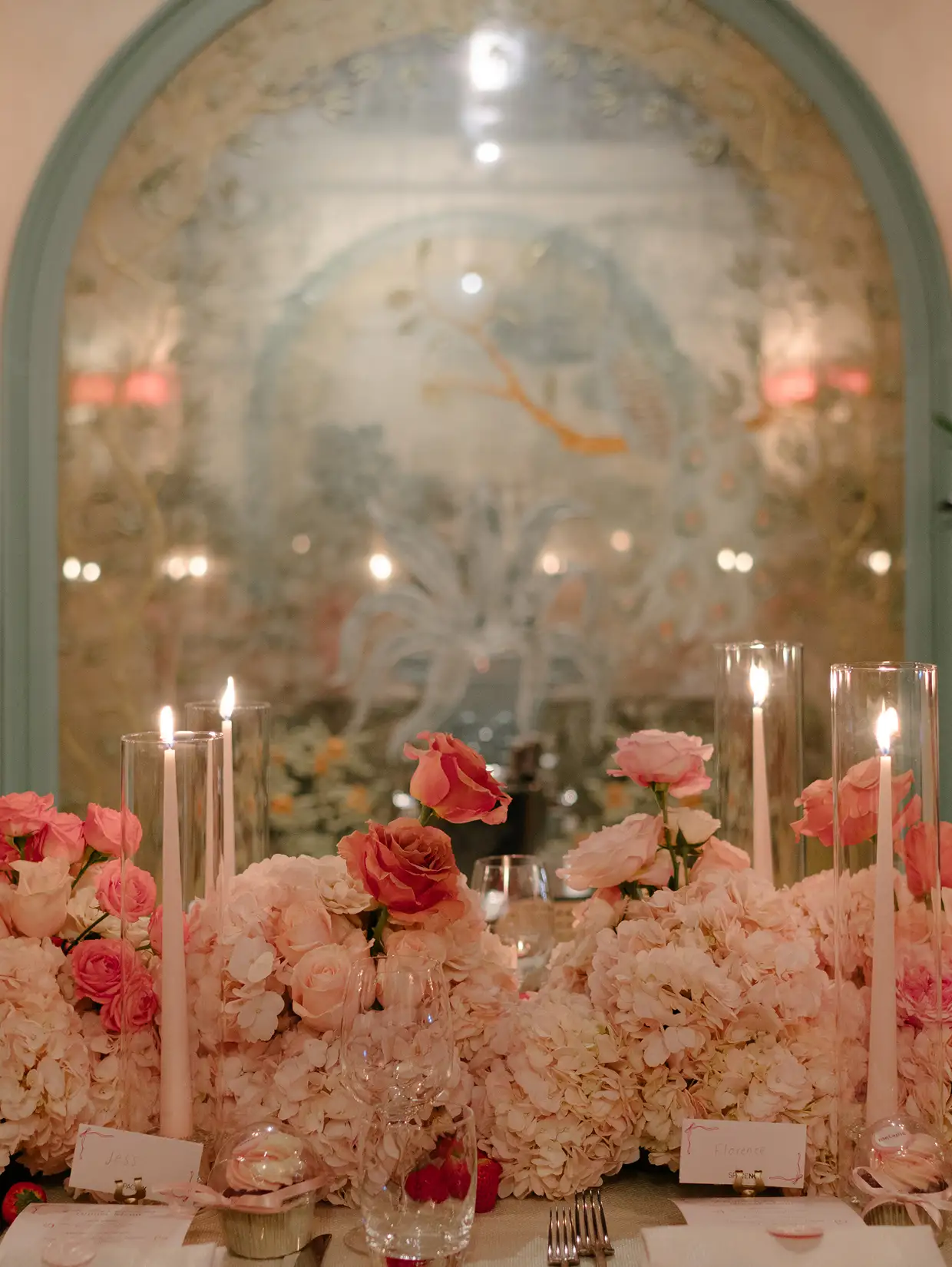 Elegant table setting with tall candles and pink floral arrangements in front of an ornate arched mirror.