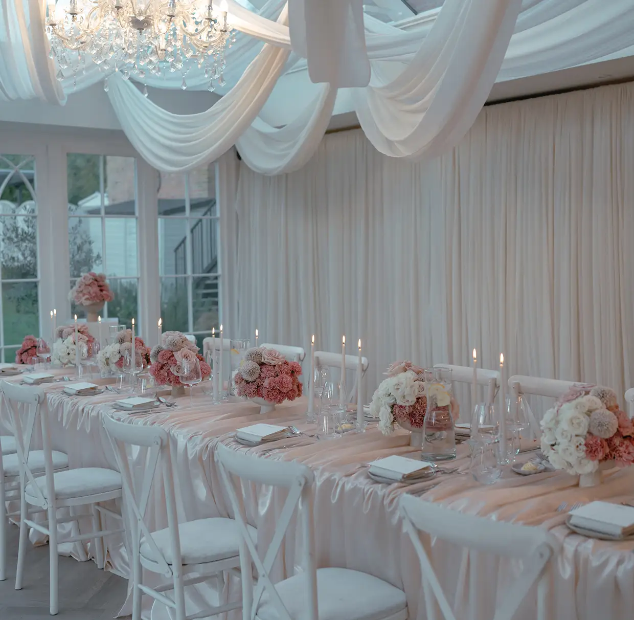 Elegant long table with white chairs, floral centerpieces, lit candles, and draped fabric ceiling in a bright room.