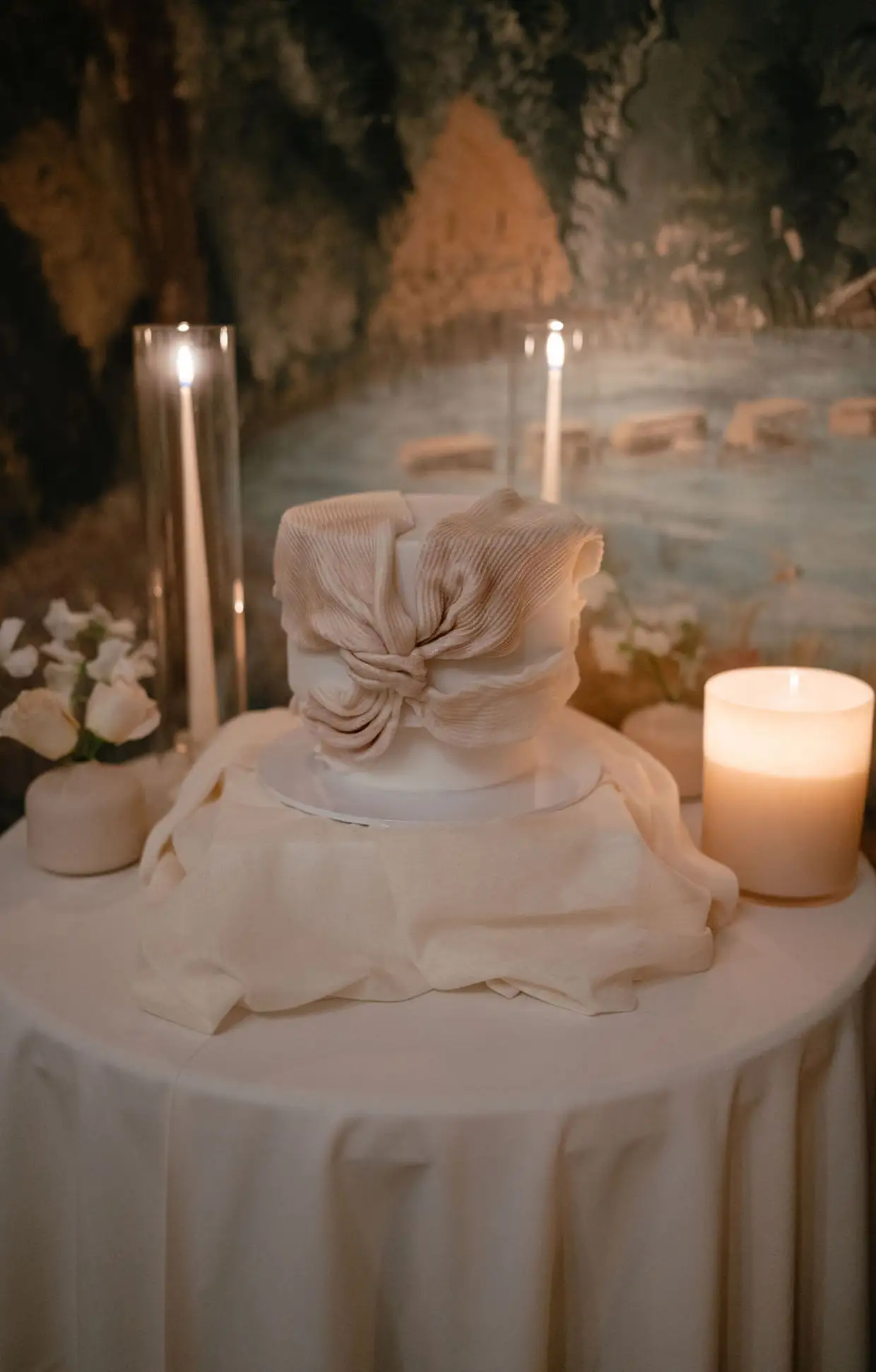 White cake with a large cream-colored bow on top, surrounded by candles and flowers on a draped round table.