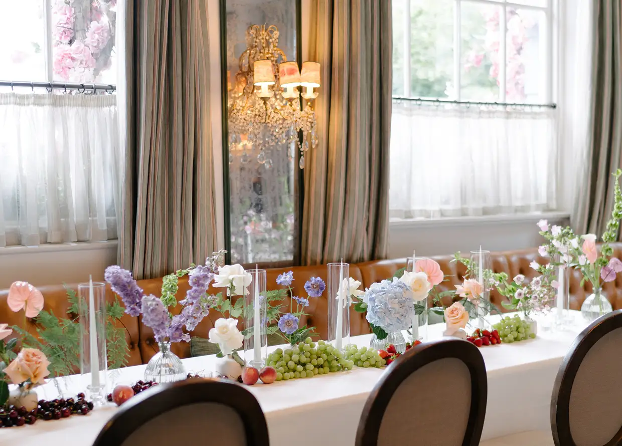 Elegant table setting with pastel flowers, candles, grapes, and fruit in a bright room with large windows.