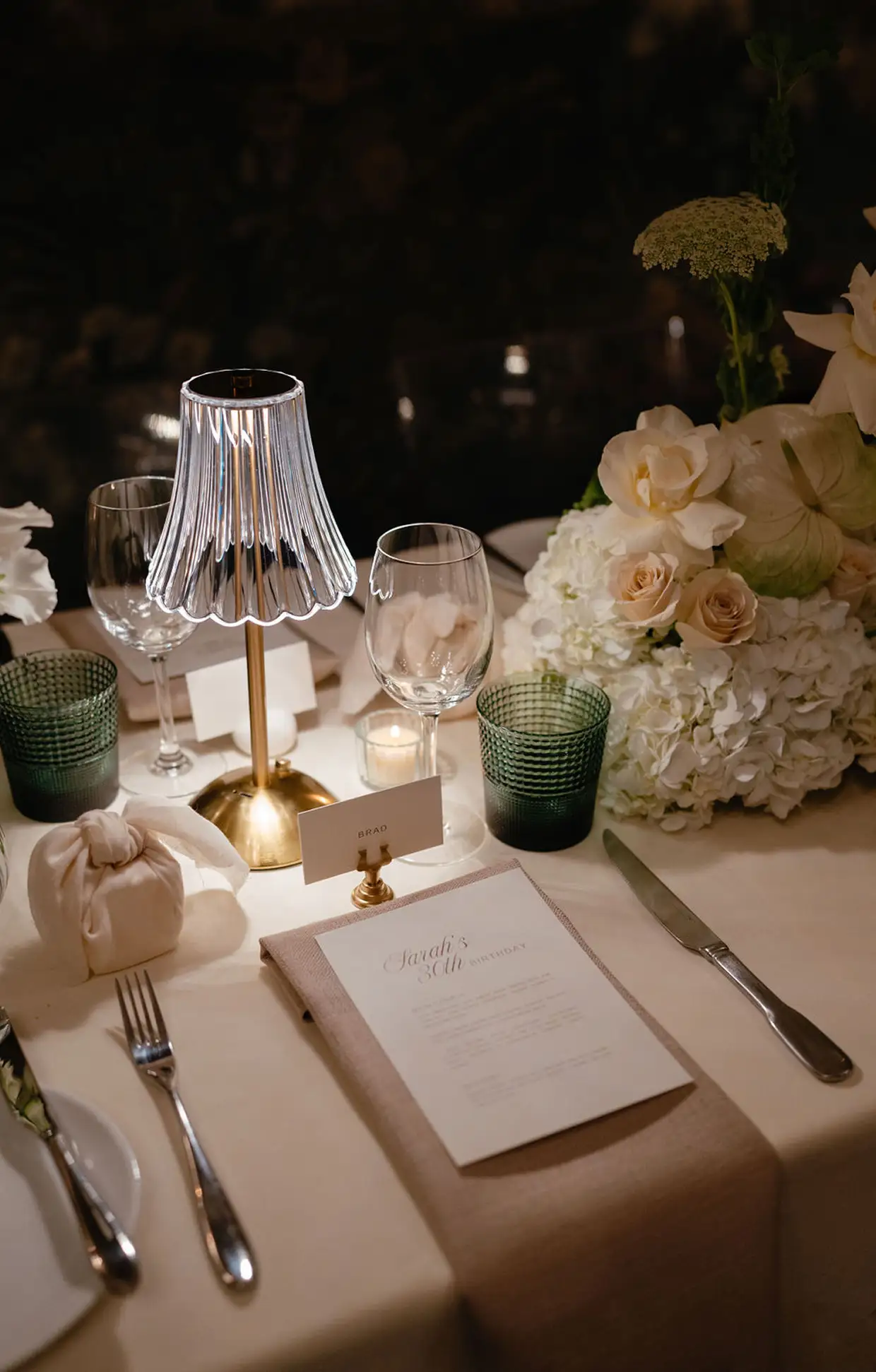 Elegant table setting with lit lamp, floral centerpiece, glassware, and a menu for Sarah's 30th birthday.