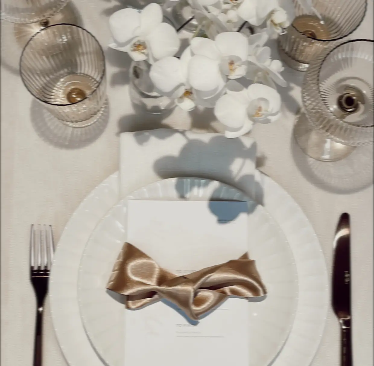 Elegant table setting with white plates, gold satin bow on menu, silver cutlery, and white orchids centerpiece.