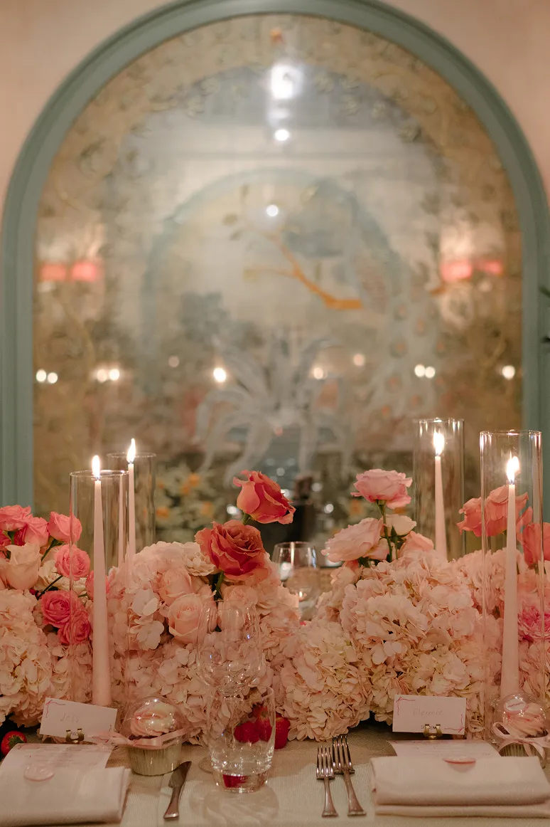 Elegant table setting with pink floral arrangements, tall lit candles, and glassware against a decorative arched backdrop.