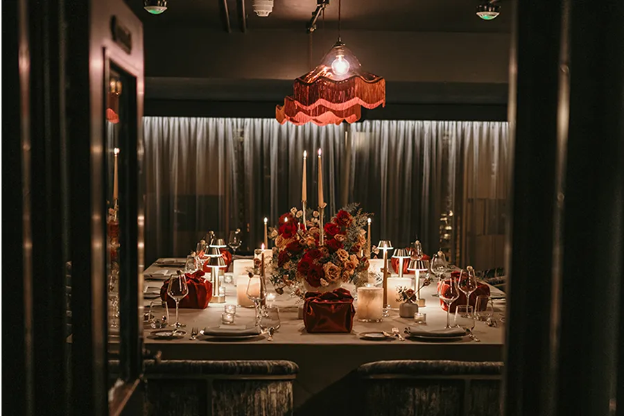 Elegant dining table set with candles, floral centerpiece, wine glasses, and red napkins under warm pendant light.