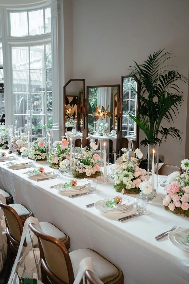 Elegantly set dining table with white tablecloth, pastel floral centerpieces, tall candles, and wooden chairs.