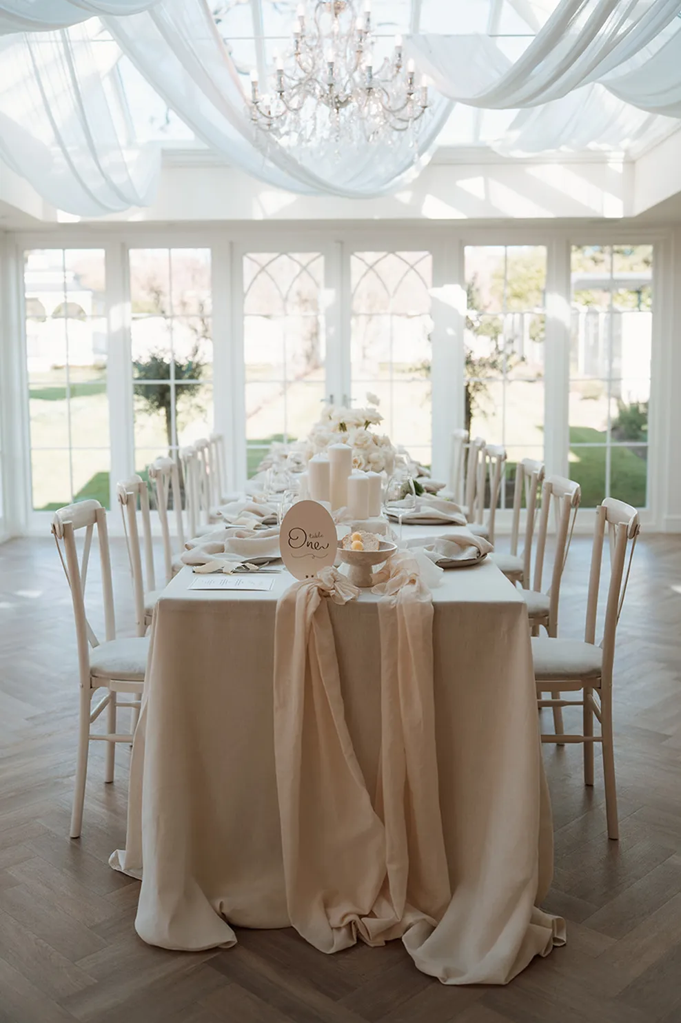 Elegant dining table with beige tablecloth, candles, flowers, and chairs in a bright room with large windows and chandelier.