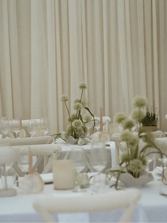 Elegant tables with white tablecloths and minimalist floral centerpieces in a neutral-toned event space.