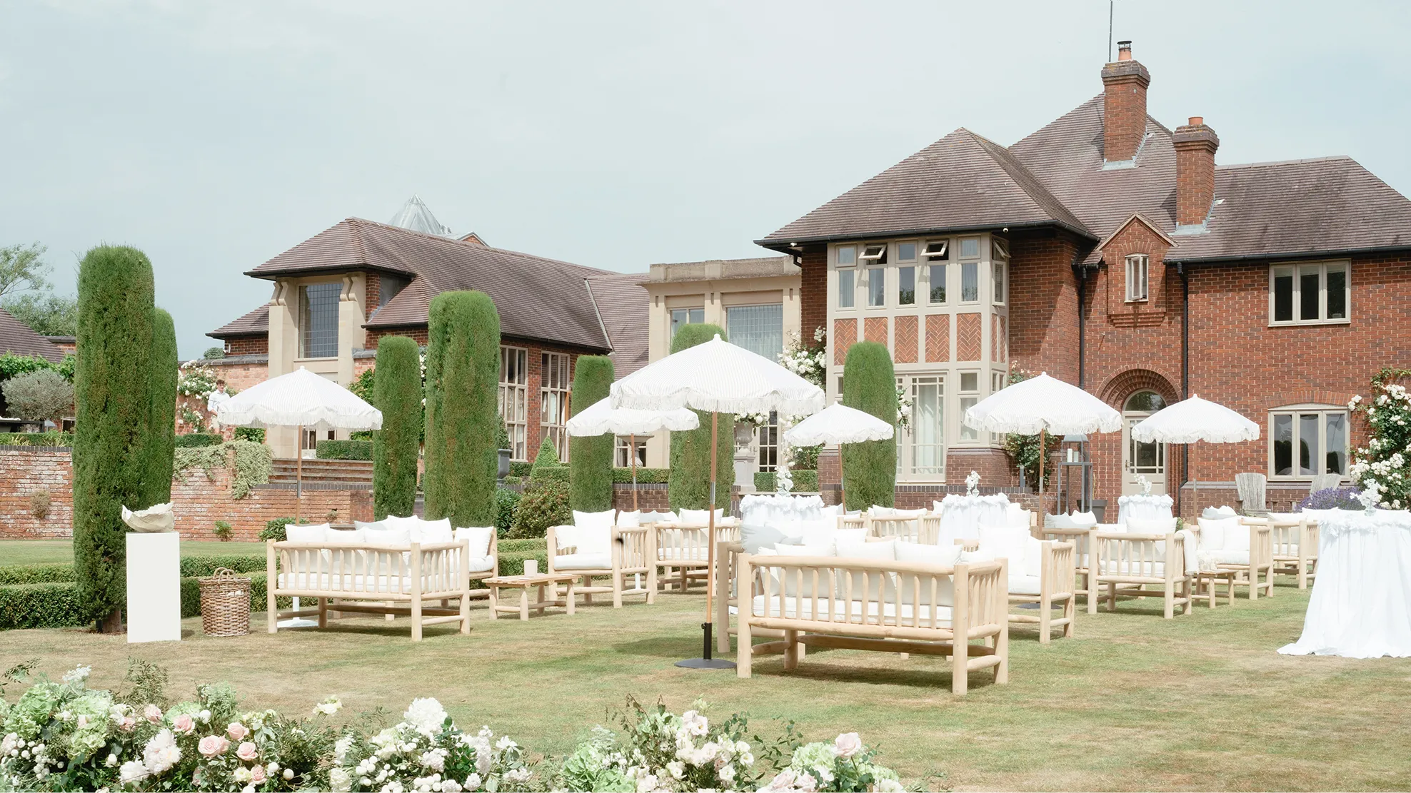 Outdoor seating area with wooden sofas and white umbrellas on a lawn in front of a brick house.