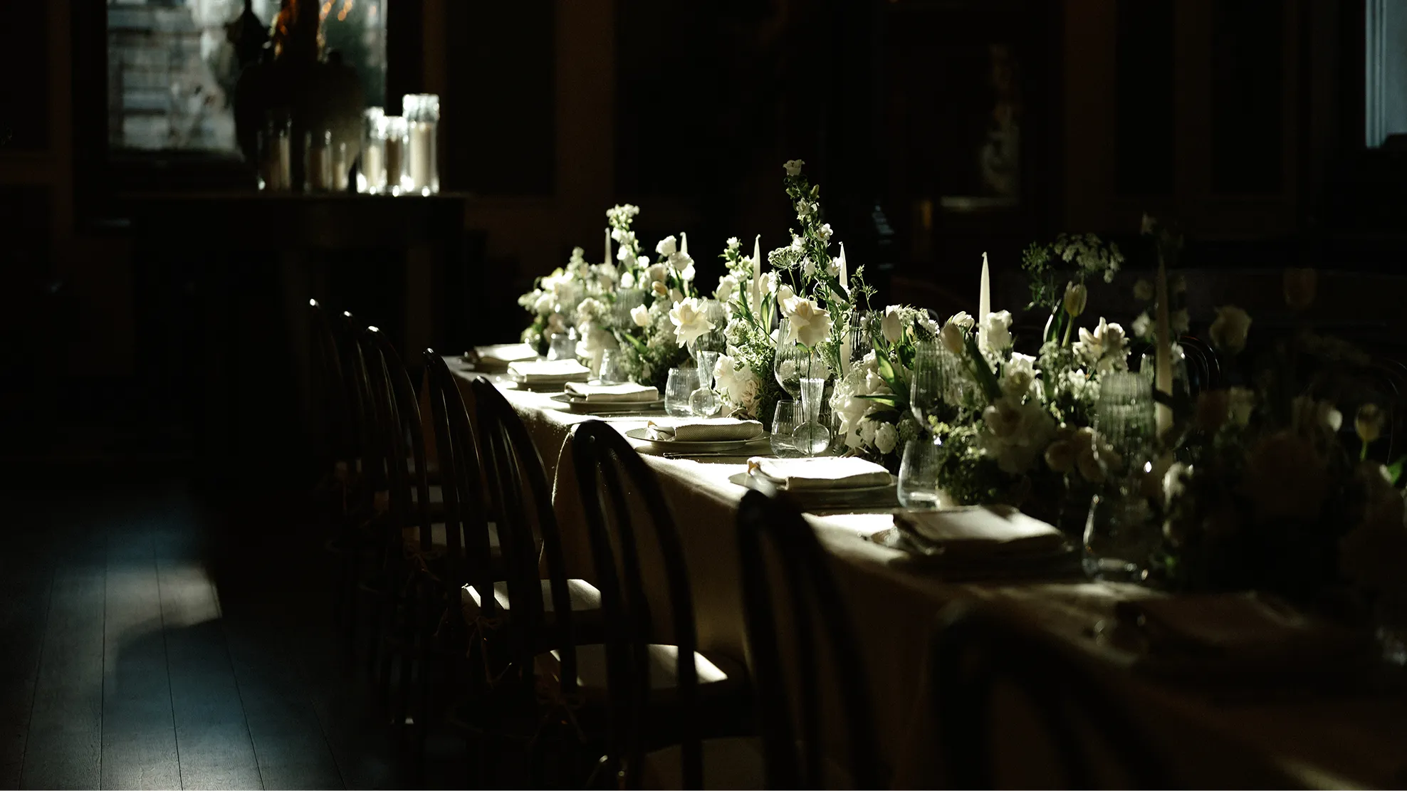 Long dining table decorated with white flowers, candles, and neatly folded napkins in dim ambient light.