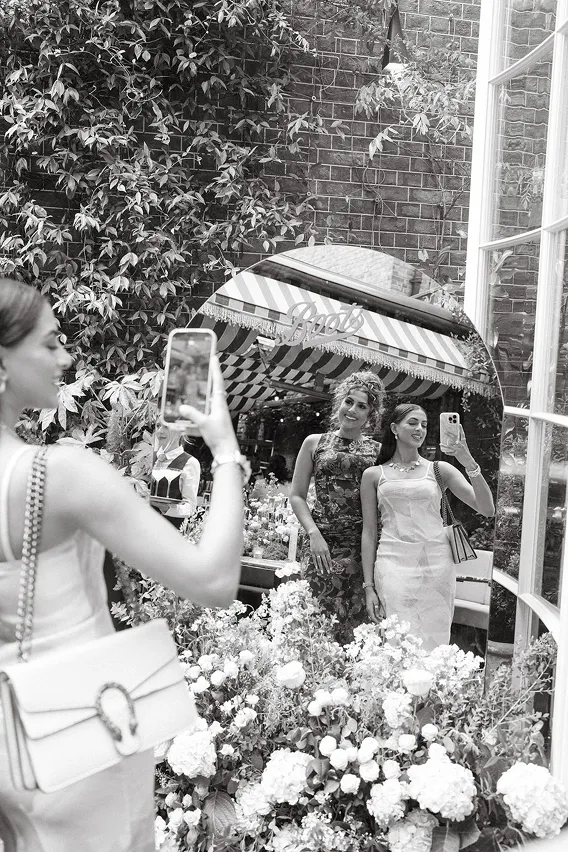Two women posing for a selfie in front of a large round mirror surrounded by flowers and greenery.