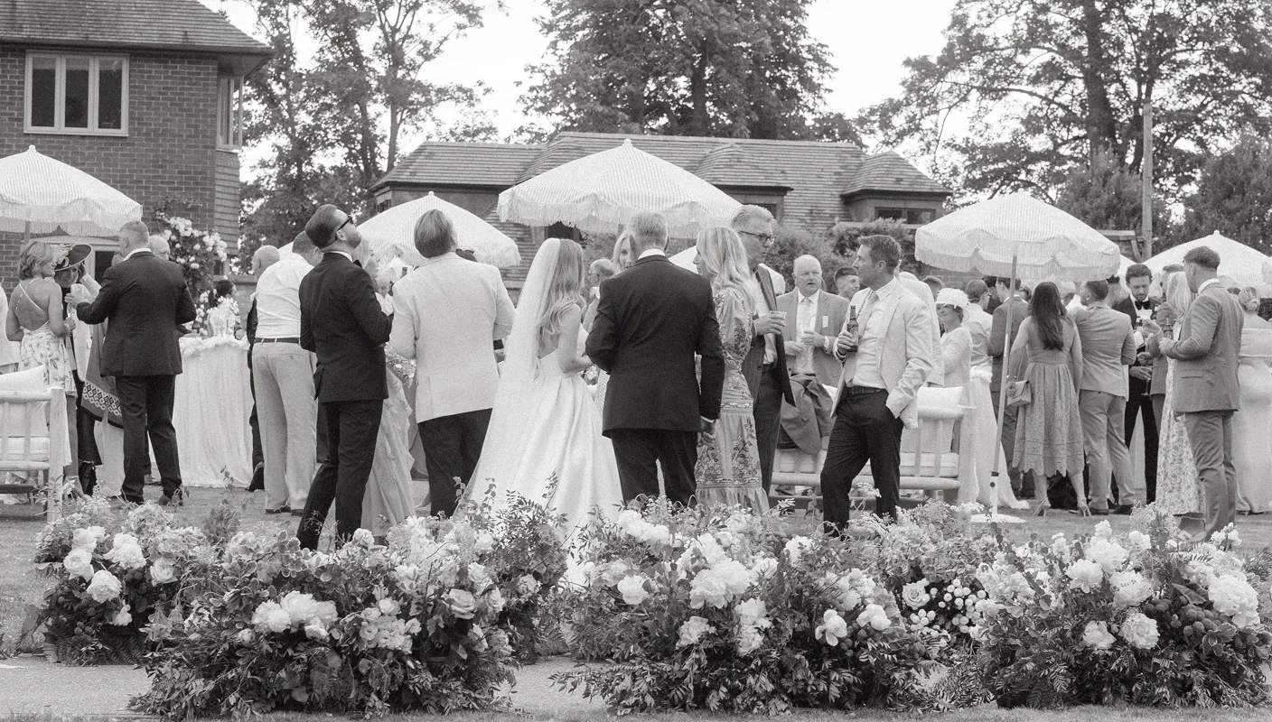 Black and white photo of a wedding reception outdoors with guests mingling and floral arrangements in foreground.