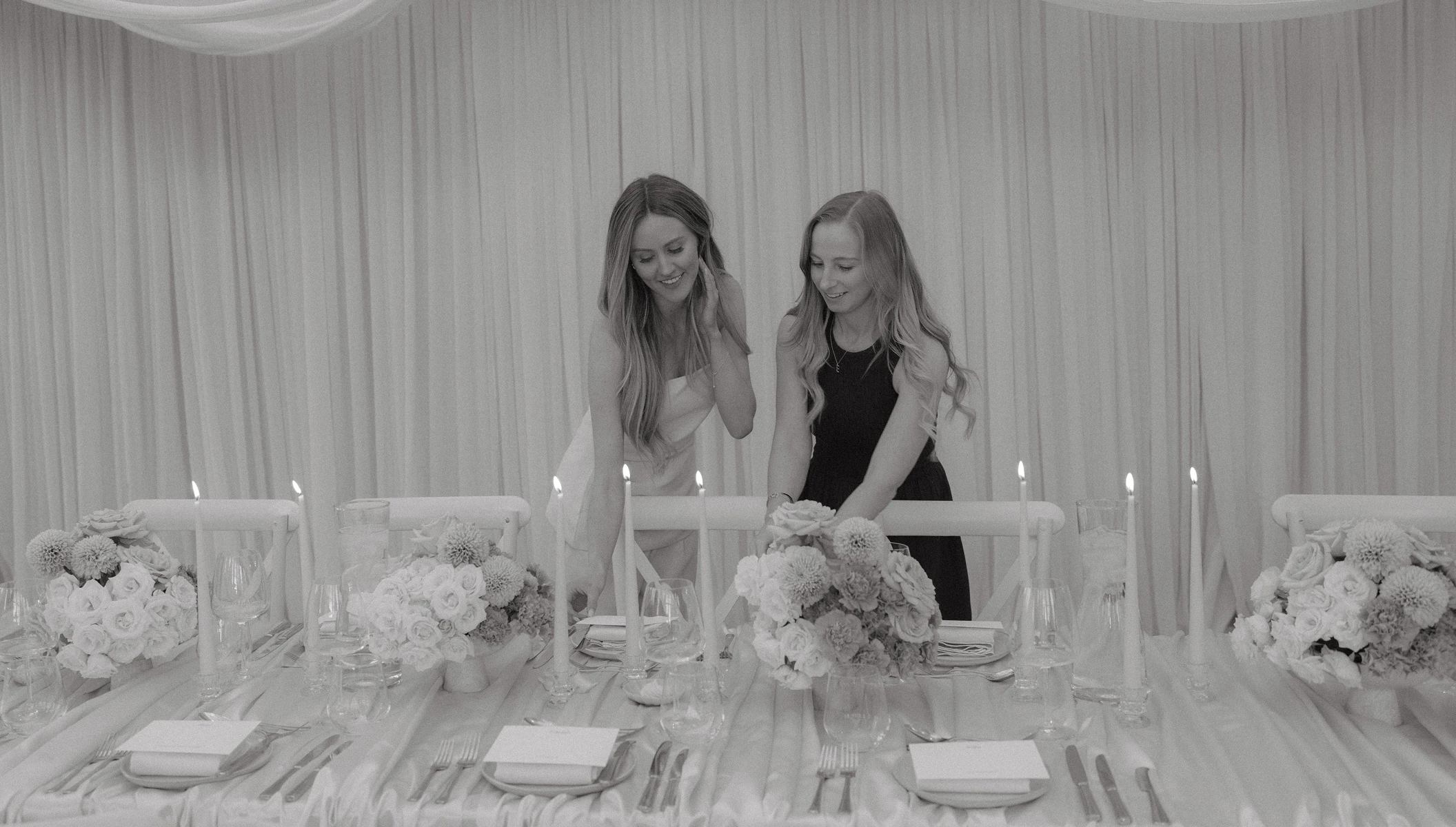 Two women smiling and arranging table settings with flowers and lit candles at a formal event.
