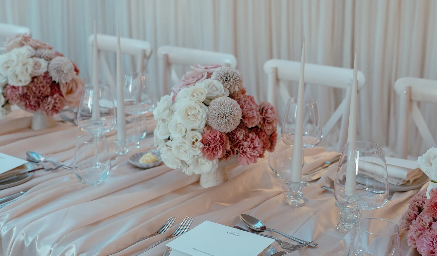 Elegant table set with pink and white floral centerpieces, candles, glassware, and silverware on a satin tablecloth.