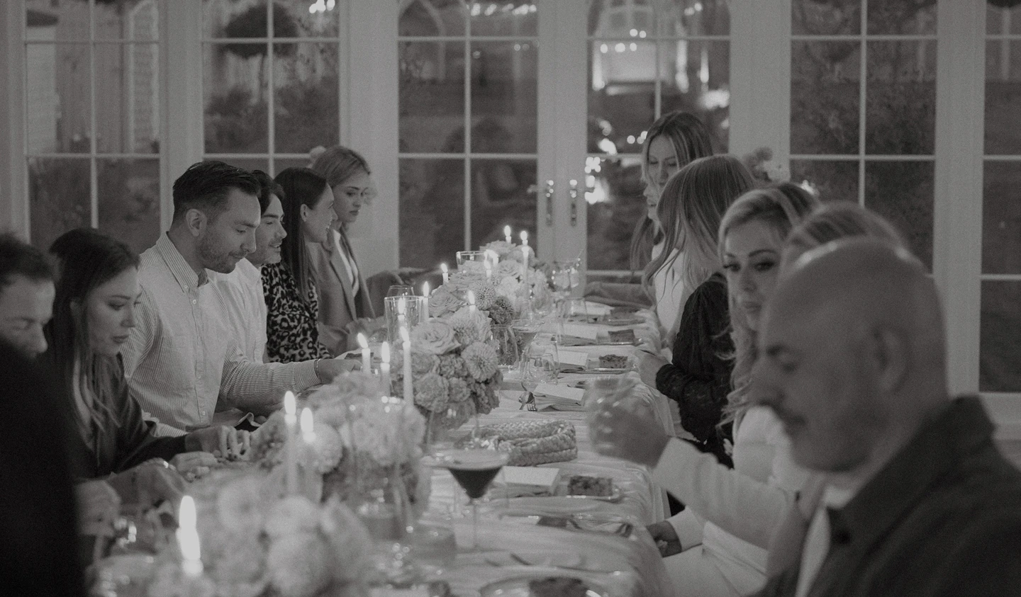 Group of people seated at a long dining table with floral centerpieces and lit candles indoors.