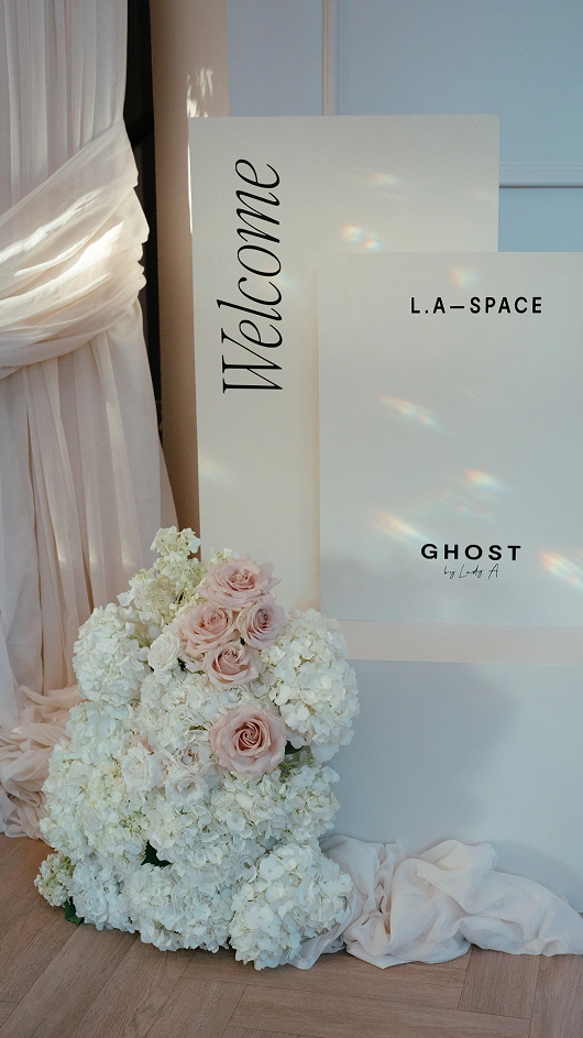 Welcome sign and L.A Space Ghost branded board next to white and pink flower arrangement by cream draped curtains.