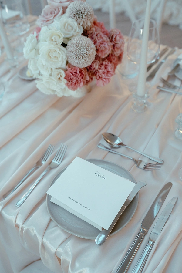 Elegant table setting with silver cutlery, a grey plate, a white folded card, and a bouquet of white and pink flowers.