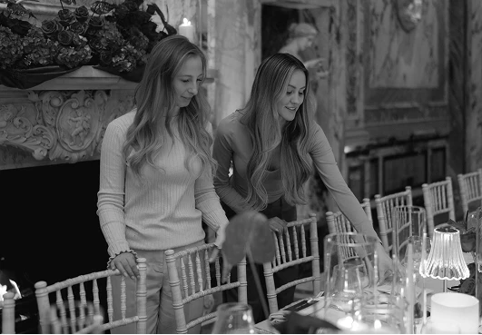 Two women setting a table in an elegant dining room with ornate decor and candles.