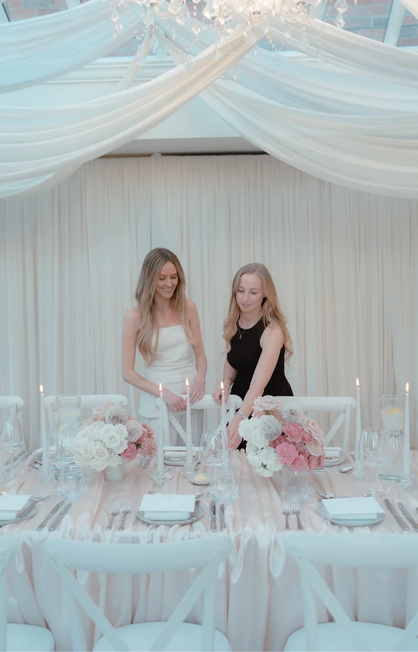 Two women smiling while arranging candles on an elegantly set dining table with flowers and draped fabric.