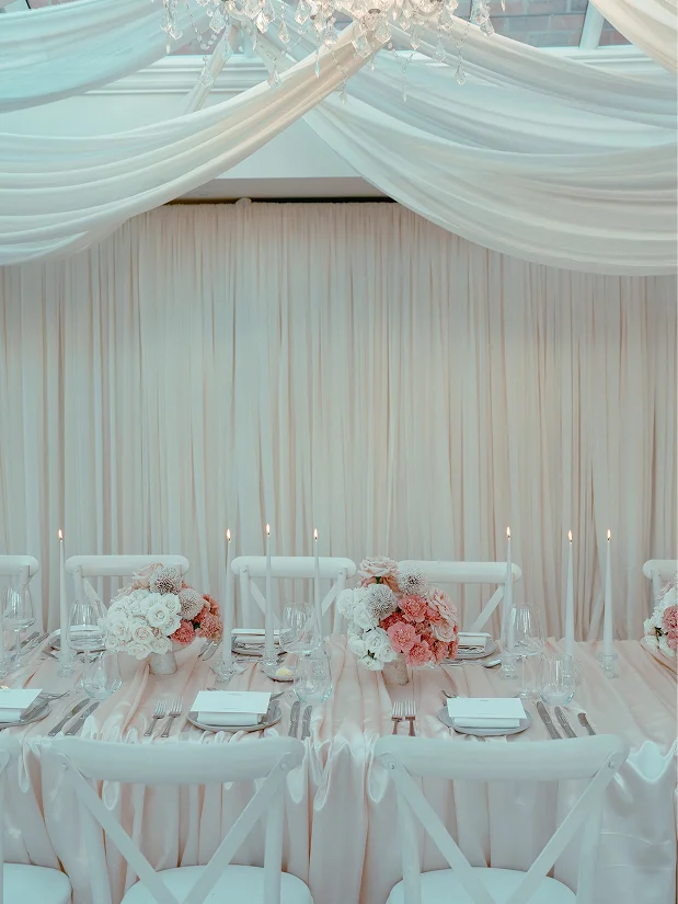 Elegant table set for event with white chairs, peach and white floral centerpieces, lit candles, and draped fabric ceiling.