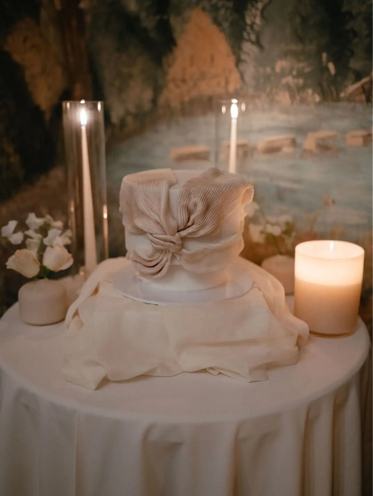 White round cake with textured fabric bow on top, on a draped table surrounded by candles and white roses.