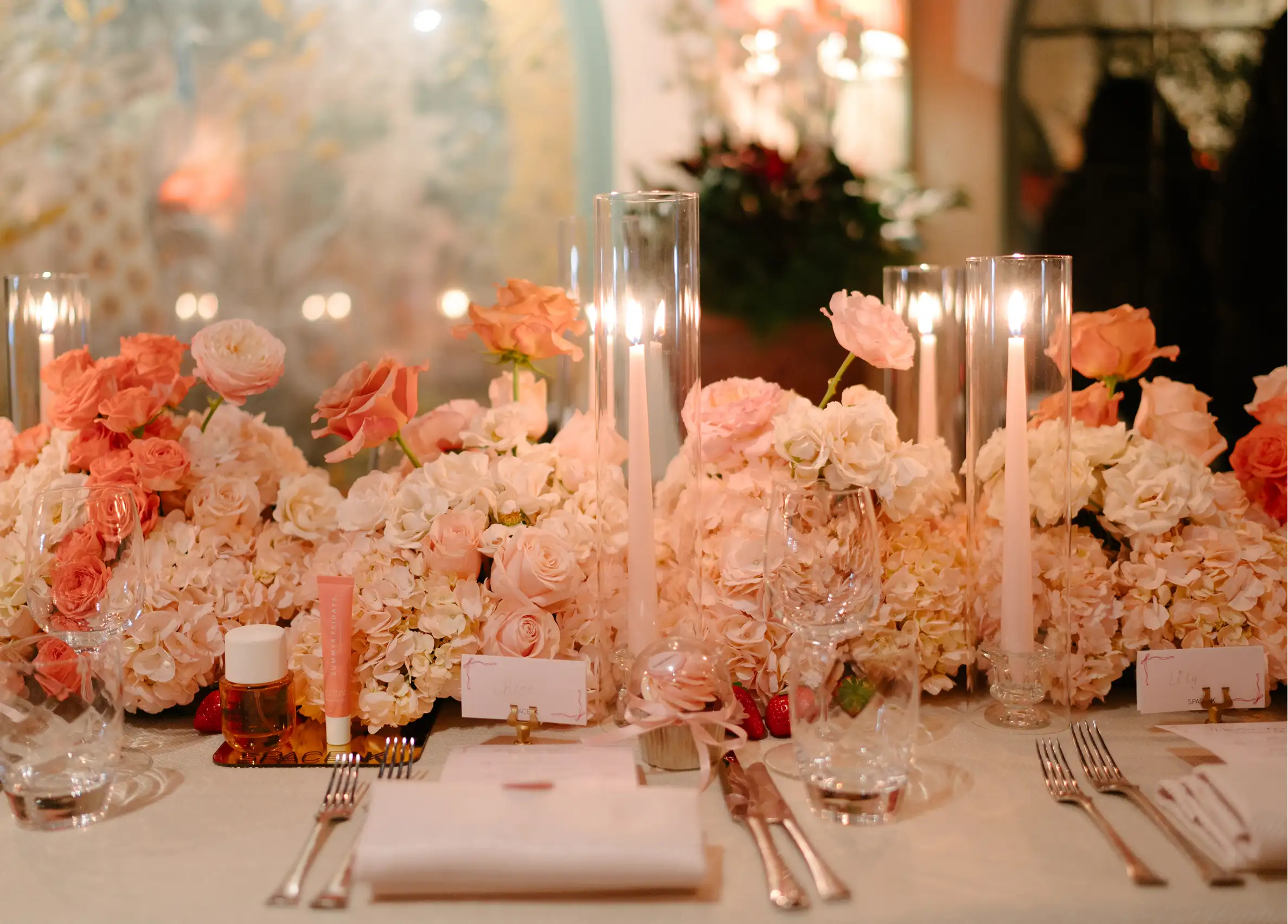 Elegant table setting with pink and peach roses, hydrangeas, glassware, and tall lit candles in glass holders.