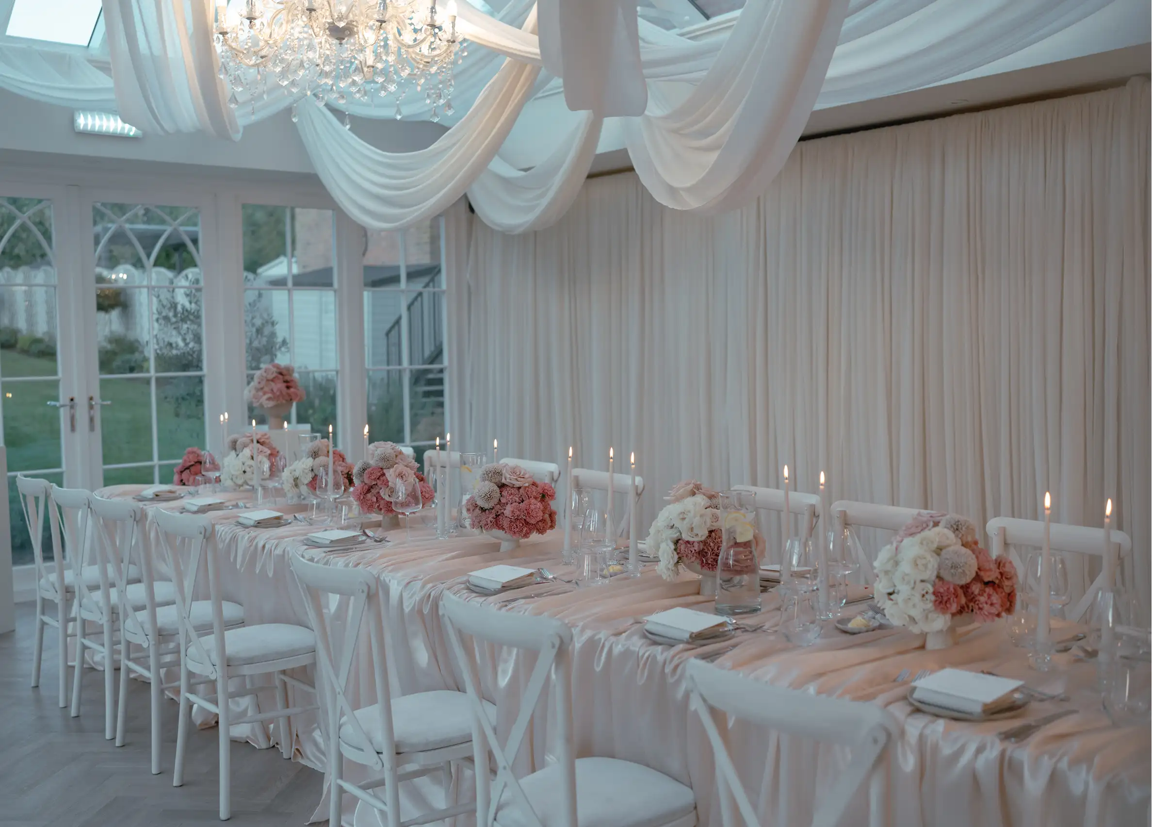 Elegant long table with white chairs, soft pink floral arrangements, lit candles, and draped fabric overhead.