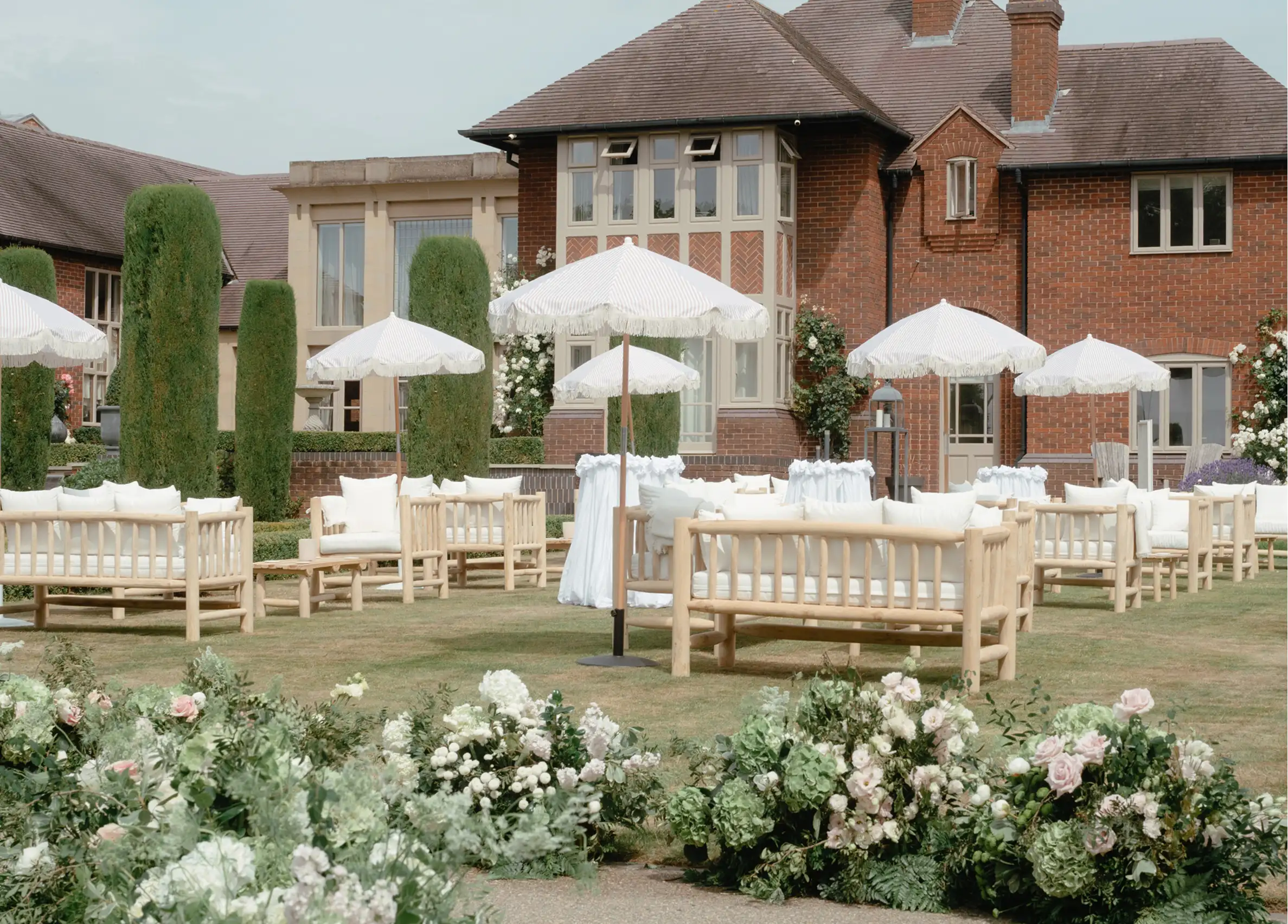 Outdoor seating area with wooden sofas, white cushions, parasols, and floral arrangements in front of a brick house.