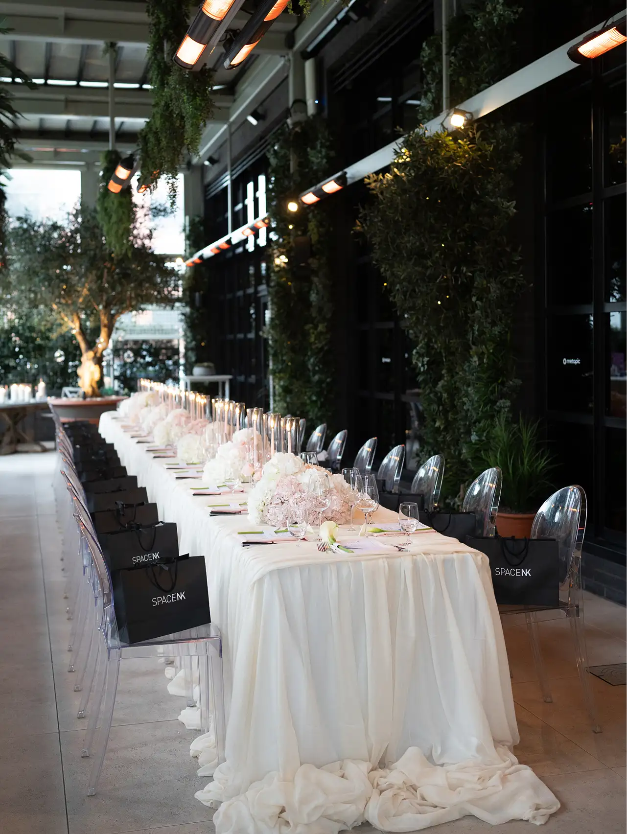 Elegant dining table with white tablecloth, floral centerpieces, candles, and transparent chairs with black SpaceNK bags.