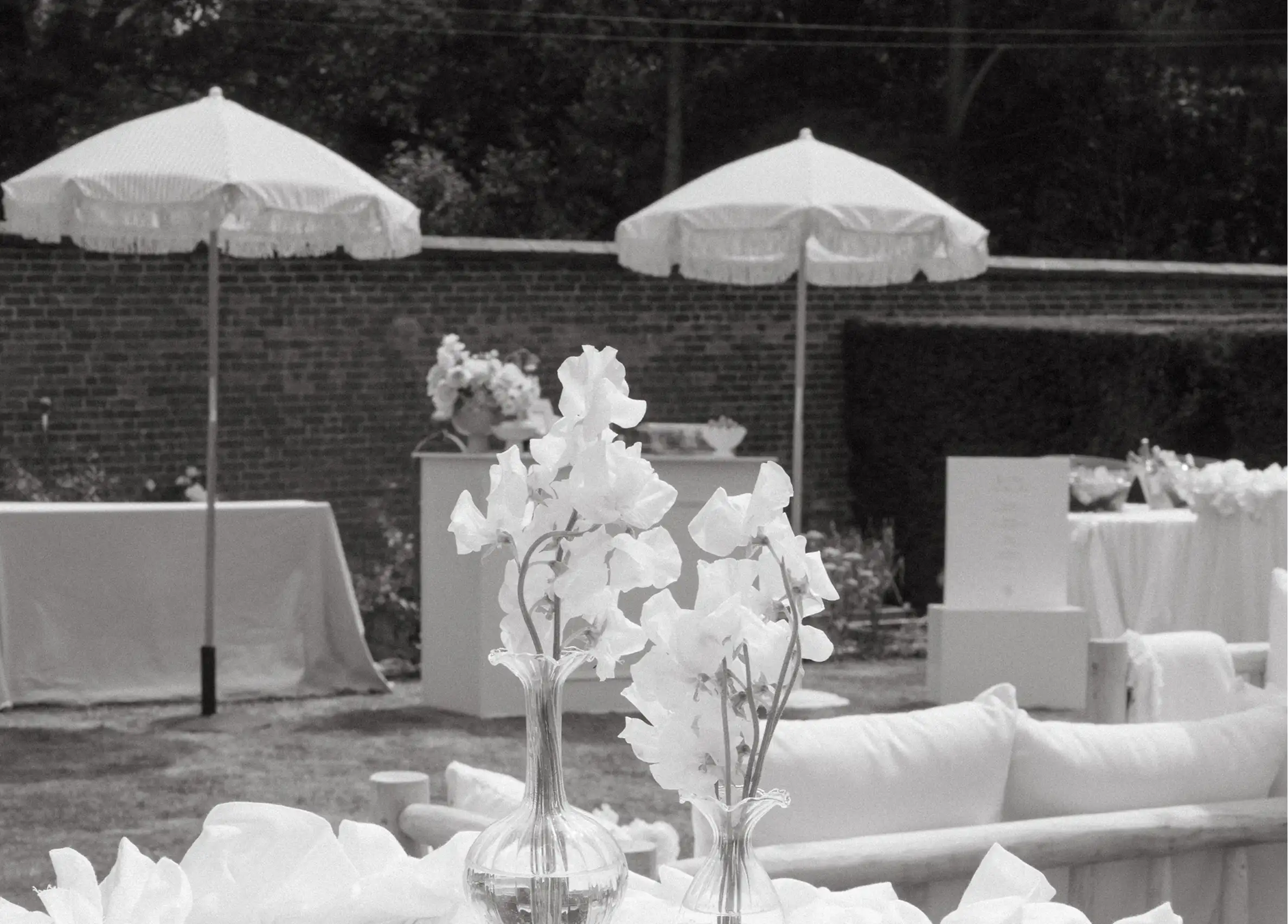 Outdoor wedding setup with white flowers in glass vases and white umbrellas against a brick wall.