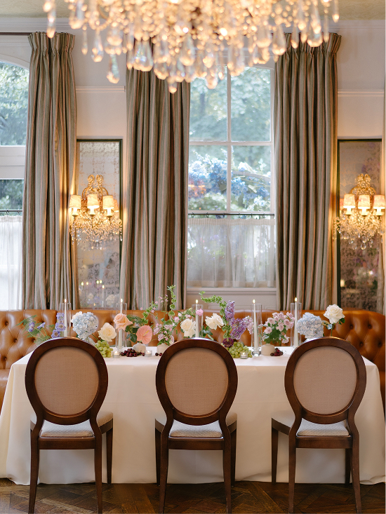 Elegant dining table set with floral arrangements and candles in front of tall windows with curtains and chandeliers.