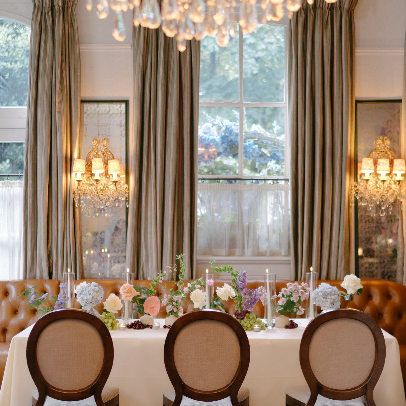 Elegant dining table with floral centerpieces and candles in a room with striped curtains and crystal wall sconces.