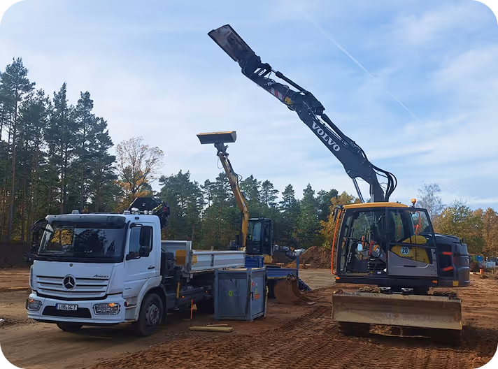 Baustelle mit weißem Mercedes-LKW und einem Volvo-Bagger mit erhobenem Arm vor bewaldetem Hintergrund unter blauem Himmel.