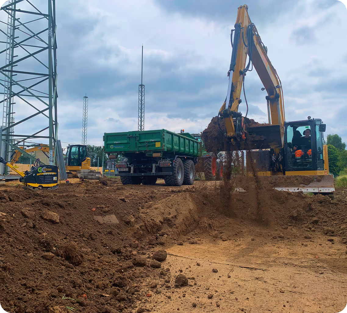 Bagger hebt Erdreich in eine Mulde auf einer Baustelle mit mehreren Maschinen und bewölktem Himmel.