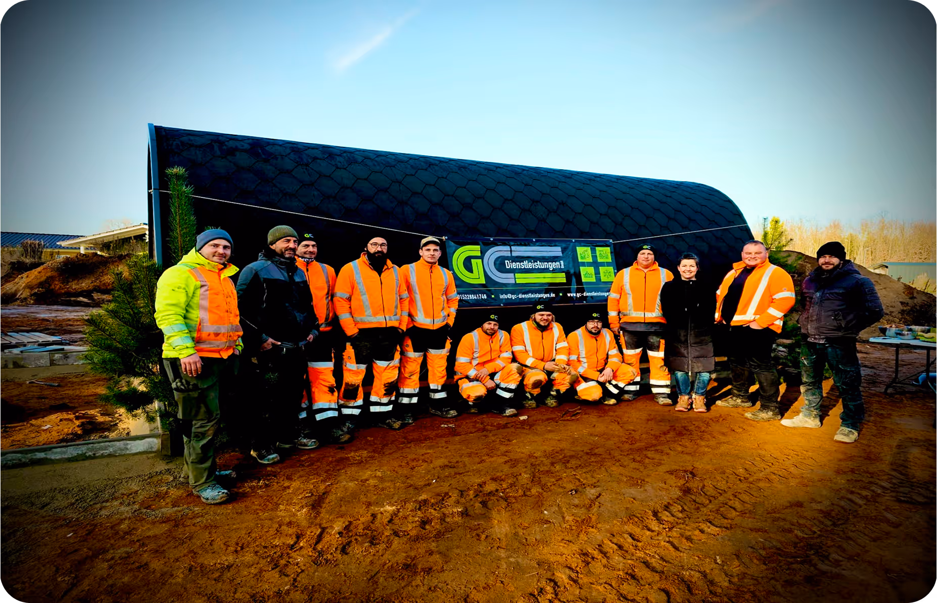 Gruppe von Bauarbeitern in orangen Warnwesten posiert vor einem schwarzen Zelt mit einem Banner bei einer Baustelle.
