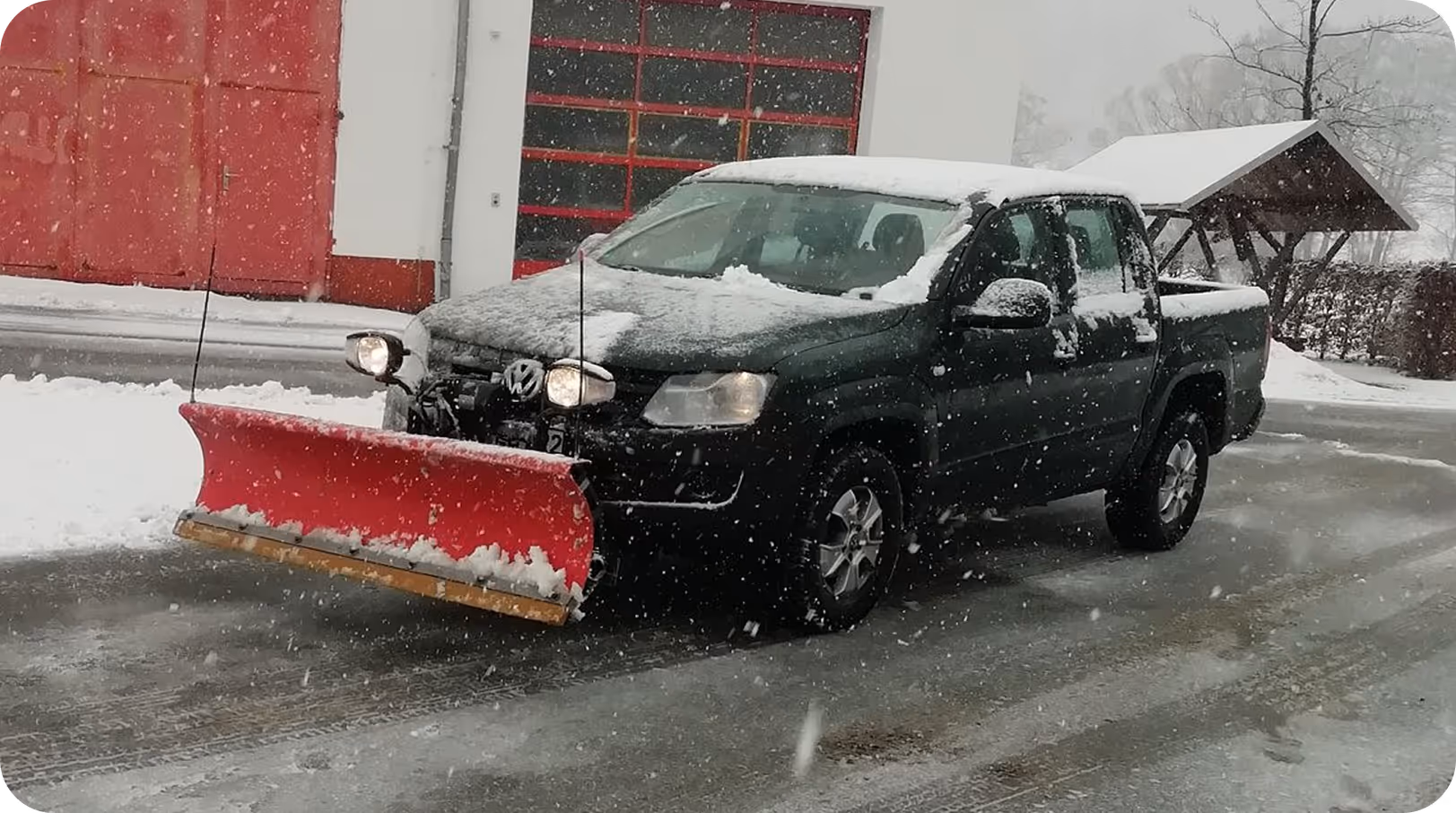 Schwarzer Pickup mit rotem Schneepflug steht auf schneebedeckter Straße vor Gebäude mit roten Türen, umgeben von Schneefall.