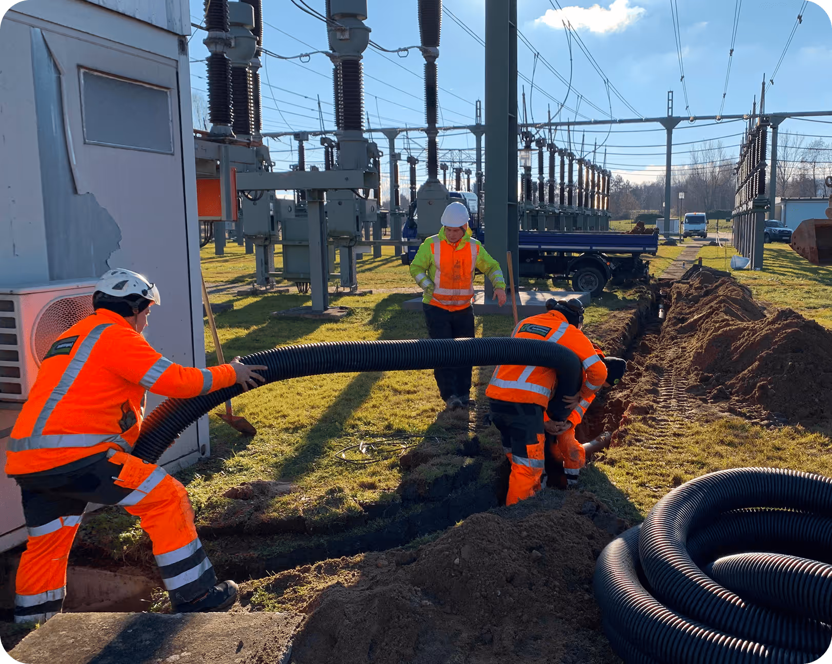 Drei Bauarbeiter in orangefarbenen Sicherheitsanzügen verlegen ein schwarzes Rohr in einem offenen Graben bei einer elektrischen Umspannstation.