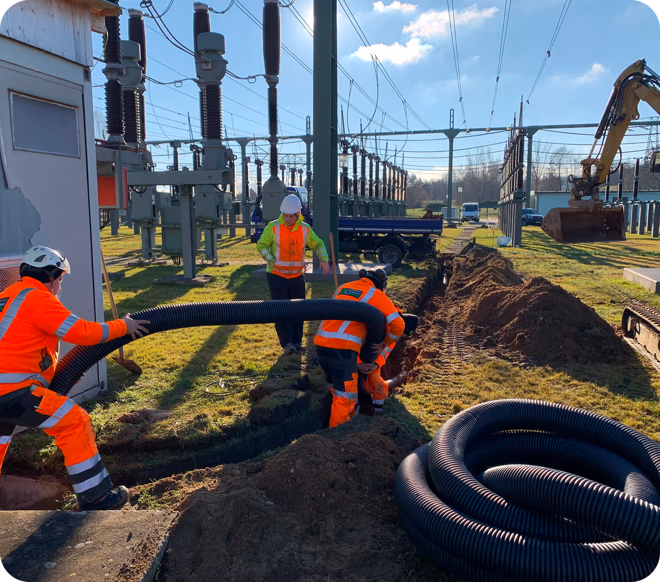 Arbeiter in orangefarbener Schutzkleidung verlegen ein große schwarze Rohrleitung in einem Kabelgraben bei einer Umspannstation.