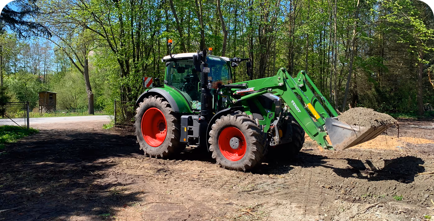 Grüner Traktor mit roten Rädern hebt eine Schaufel voll Erde auf einem bewaldeten Feld.