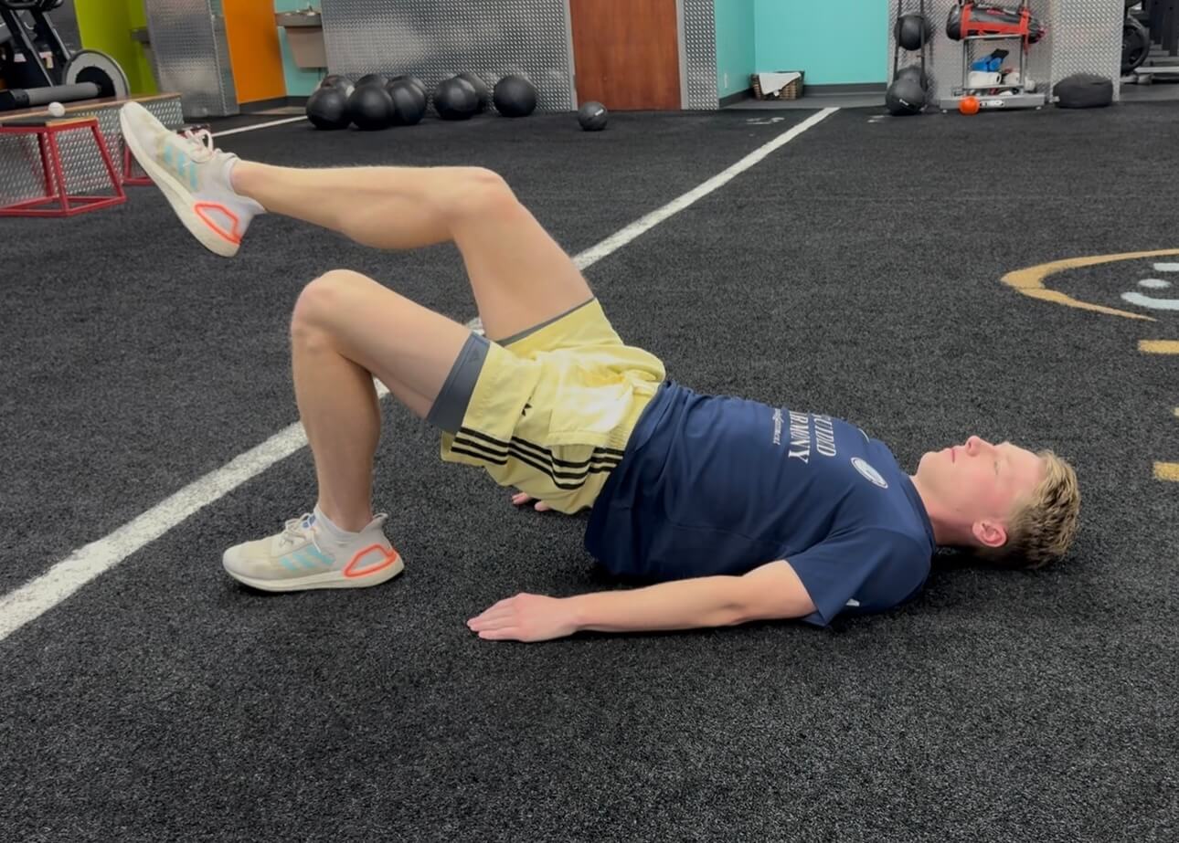 Person lying on their back on gym floor performing single-leg bridge exercise with one leg raised.