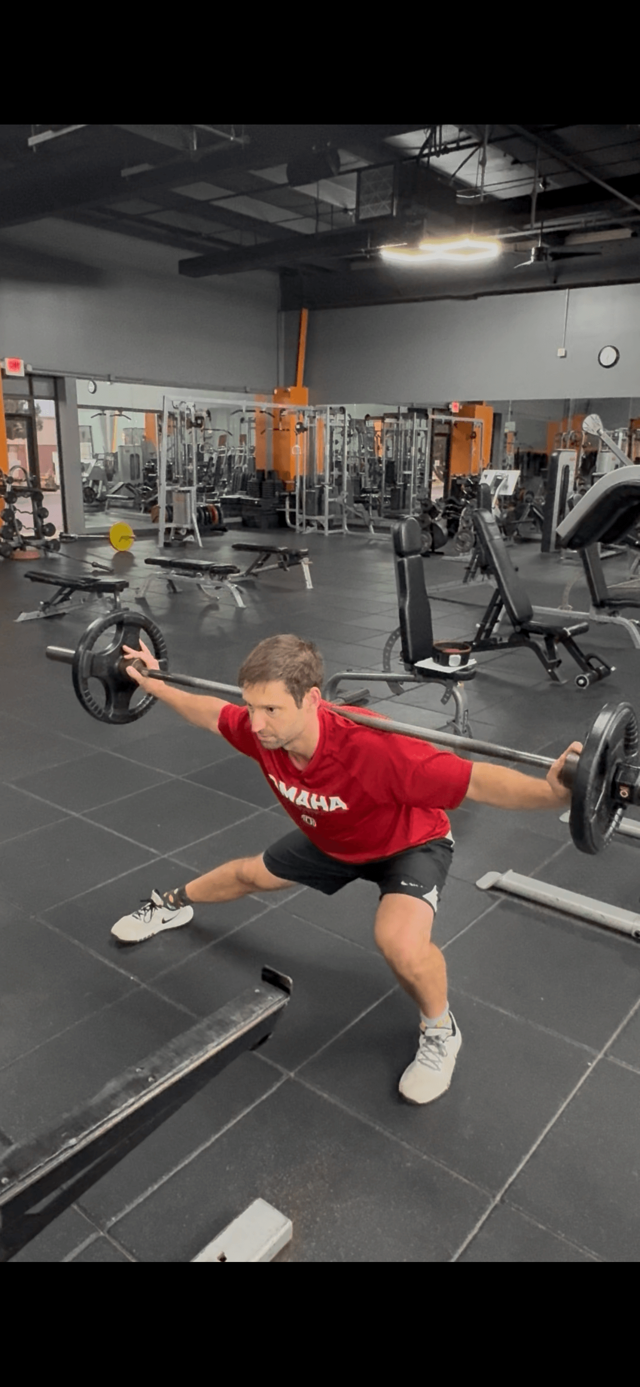 Man in a red shirt performing a wide squat with a barbell on his shoulders in a gym.