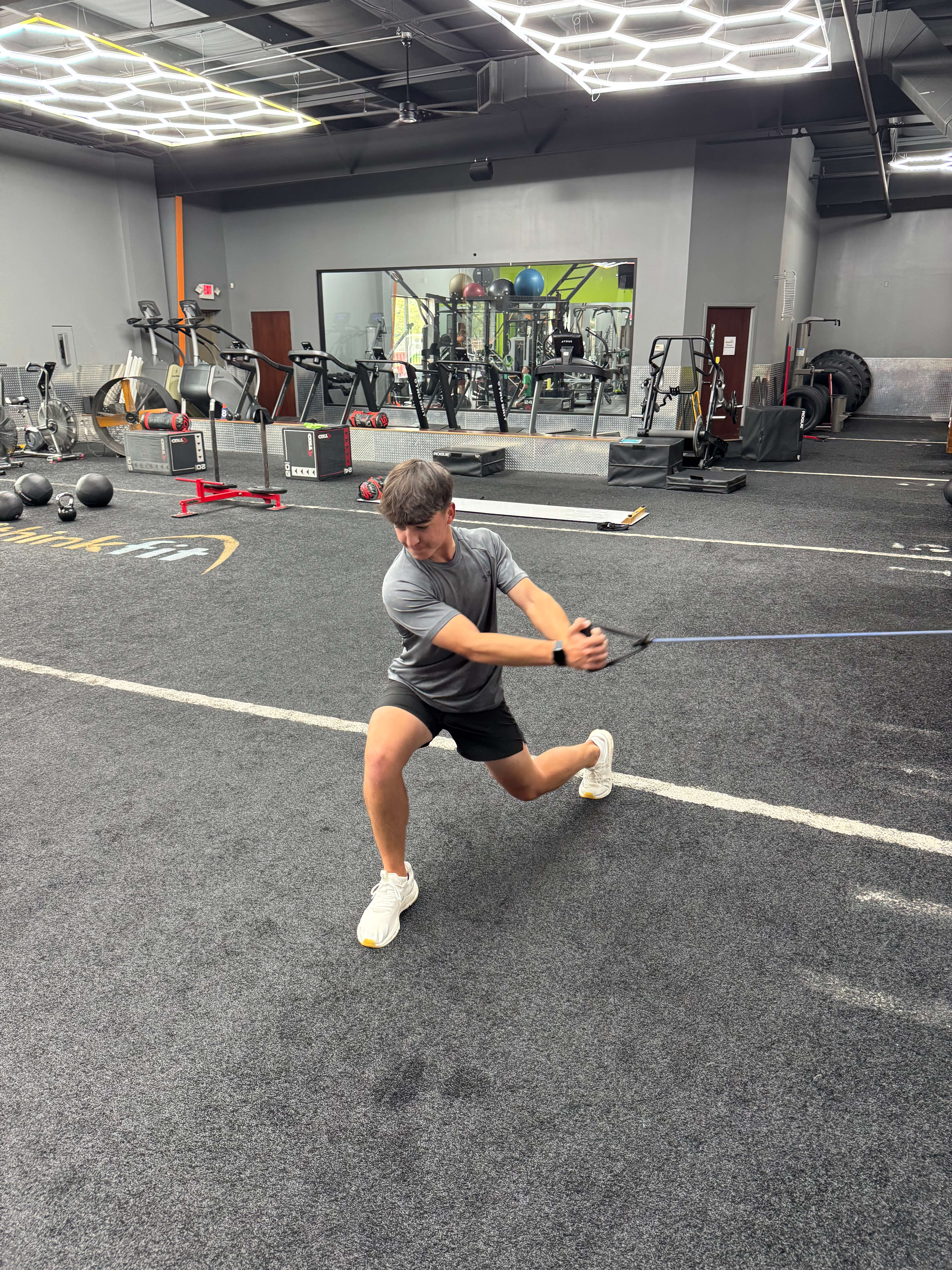 Young man in gray shirt and black shorts performing a resistance band exercise in a gym with exercise equipment and mirrored wall in the background.
